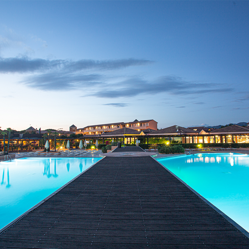 Un camminamento di legno si estende verso la piscina del Garden Toscana Resort, illuminato dalla luce calda del tramonto.